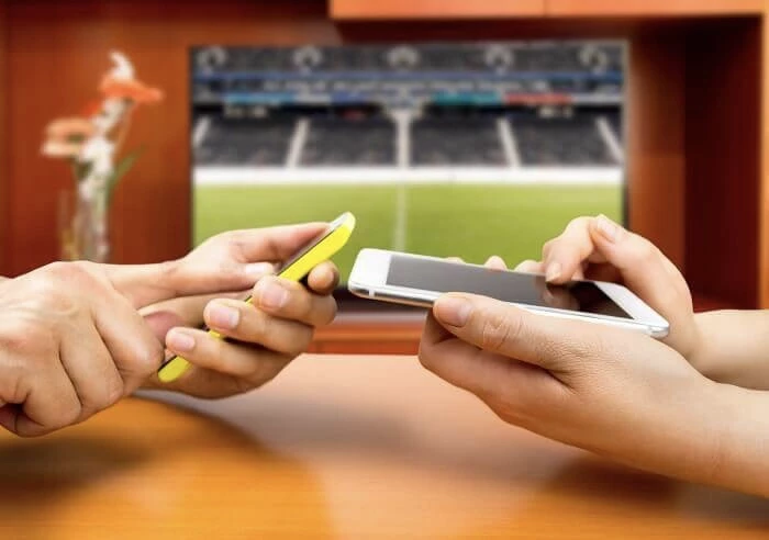 Duas mão em cima de uma mesa, cada uma a segurar um telemóvel, com uma televisão de fundo ligada a transmitir um jogo de futebol